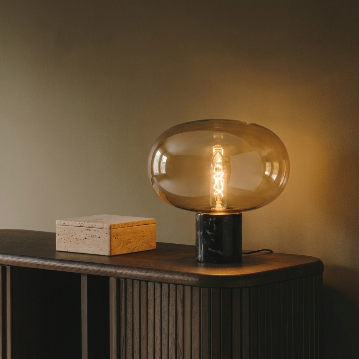 Lampe à poser en verre ambré et base en marbre noir, ampoule à filament allumée diffusant une lumière chaude, posée sur une enfilade en chêne cannelé foncé, boîte en travertin à côté