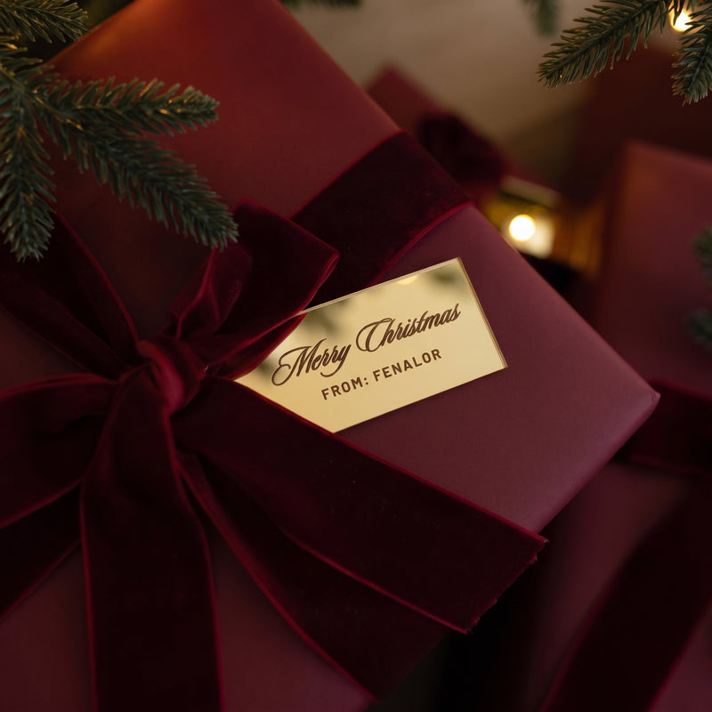 Paquet cadeau rouge noué d'un ruban de velours bordeaux, étiquette dorée Merry Christmas, sous un sapin