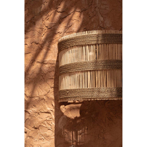 Abat-jour Doedoe allumé sur un mur enduit de terre avec la lumière qui passe entre les tiges de rotin