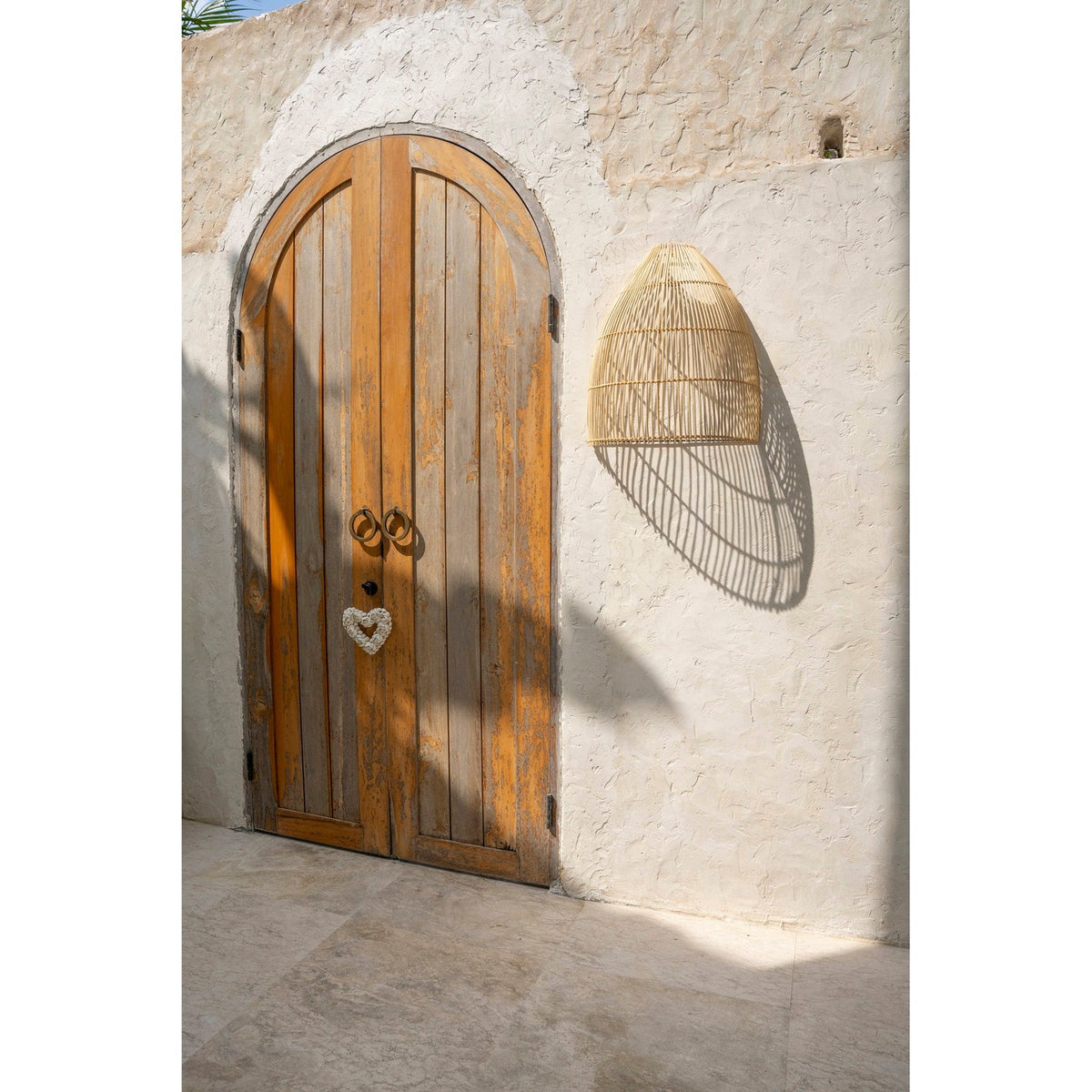Abat-jour Lalo en rotin naturel fixé sur un mur blanc à côté d'une porte cintrée en bois avec son ombre