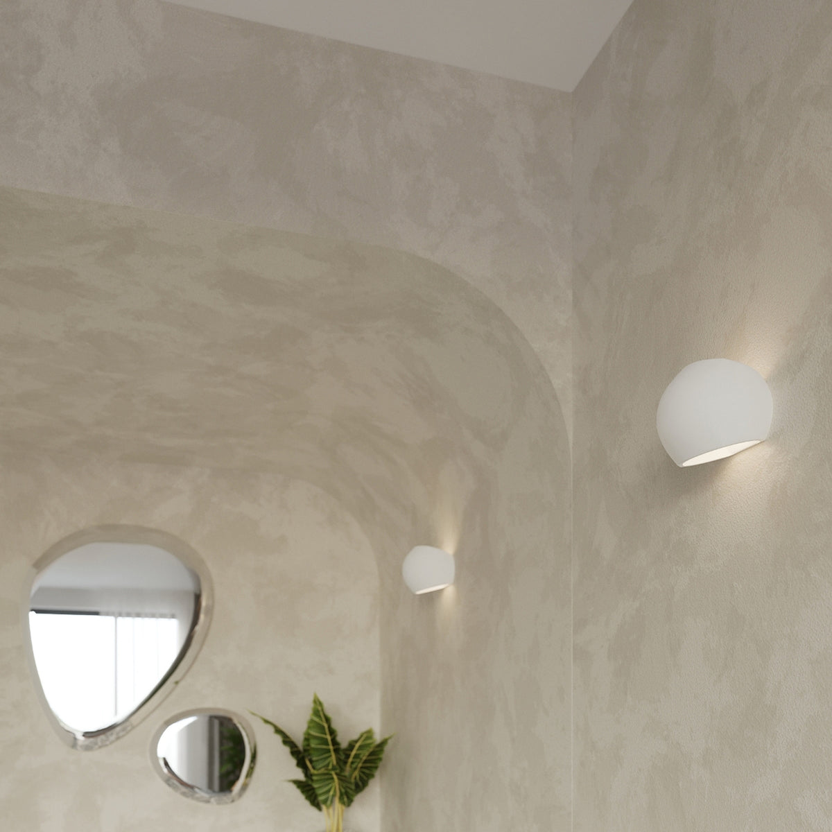 Deux appliques murales Globe blanches allumées sur un mur enduit près de miroirs organiques et d'une plante