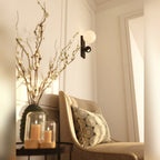 Applique Atom noire allumée au-dessus d'un fauteuil beige et d'un vase de branches fleuries