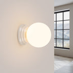 Applique murale Zori en céramique émaillée blanche allumée sur un mur clair avec sa boule qui diffuse une lumière chaude