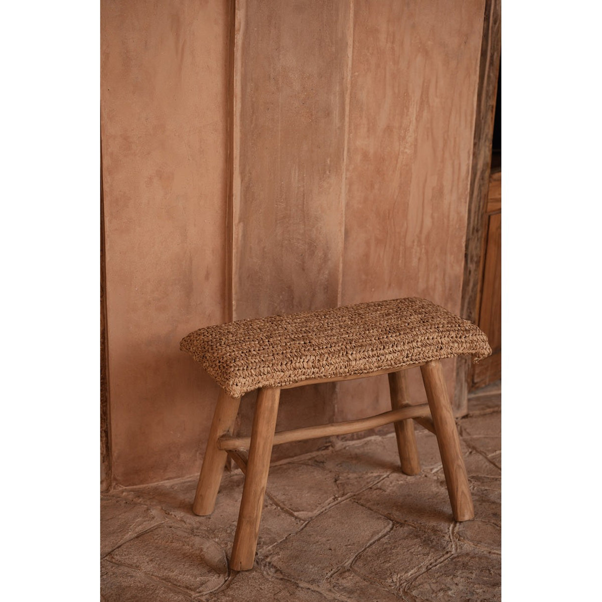 Banc Raffia en raphia et teck de trois quarts devant un mur en enduit terracotta sur un sol en pierre