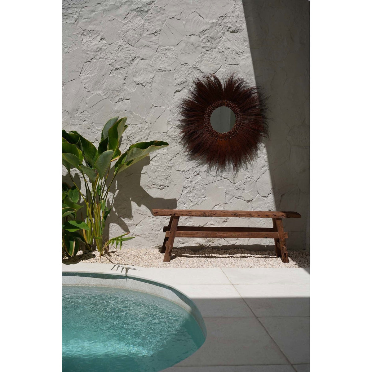 Banc Beauteous en teck contre un mur blanc sous un miroir en fibres naturelles près d'une piscine