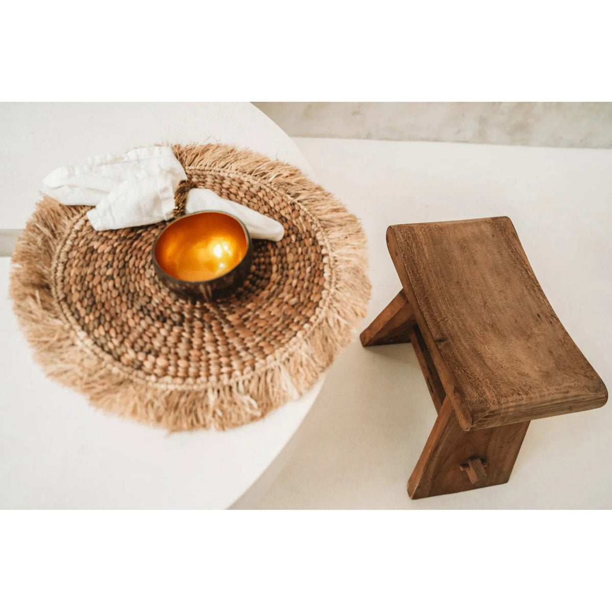 Bol en coco doré sur un set de table rond à franges avec une serviette en lin près d'un tabouret en bois