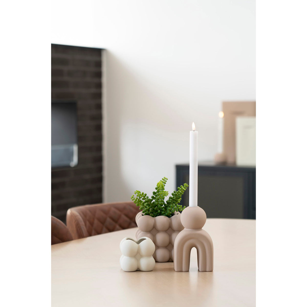 Bougeoir Arche sable avec une chandelle allumée sur une table en bois clair près de pots à bulles et d'une plante