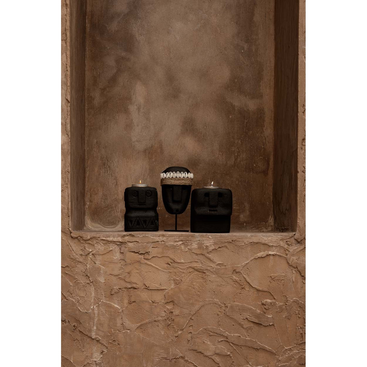 Bougeoir visage en pierre noire allumé dans une niche à côté d'un masque sur socle et d'un autre bougeoir