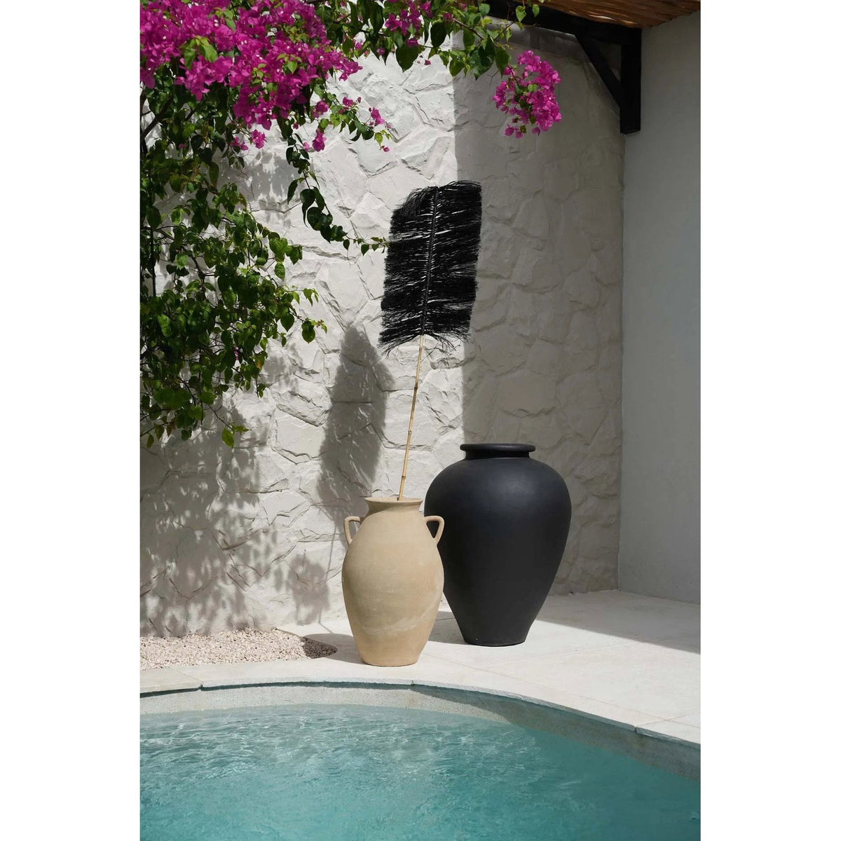 Branche Palmeira noire dans une amphore beige à côté d'une grande jarre noire au bord d'une piscine sous un bougainvillier