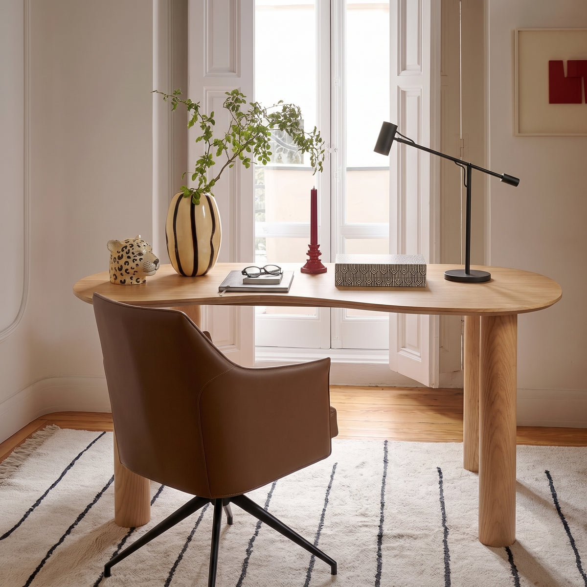 Bureau Livigno devant une fenêtre, avec un fauteuil pivotant brun, une lampe articulée noire et un vase rayé sur le plateau