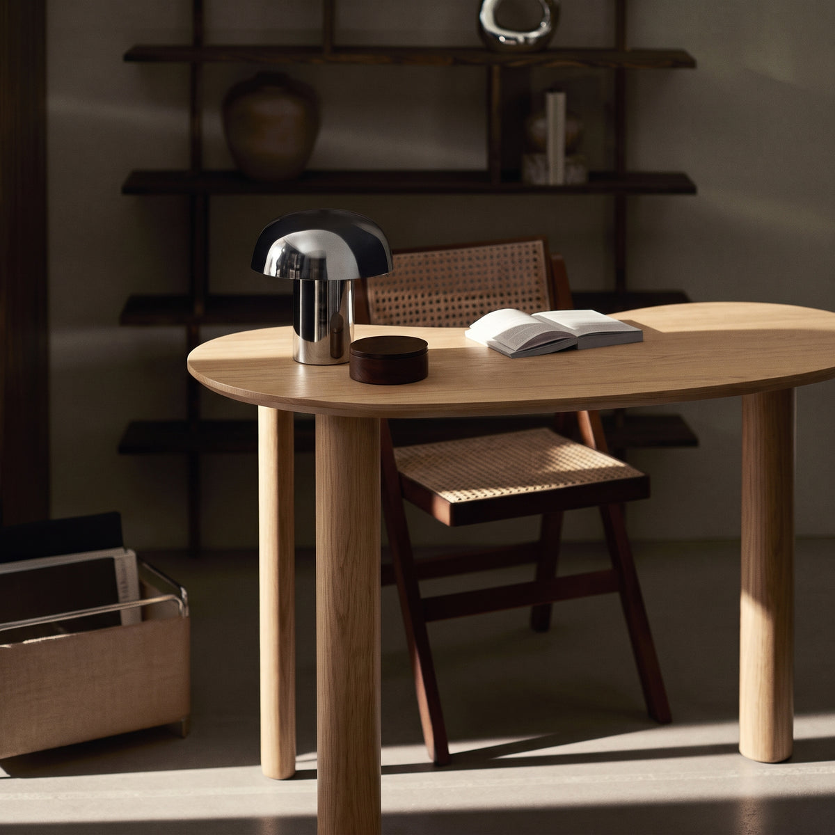 Bureau Livigno dans une lumière rasante, avec une chaise cannée, une lampe champignon chromée et un livre ouvert sur le plateau