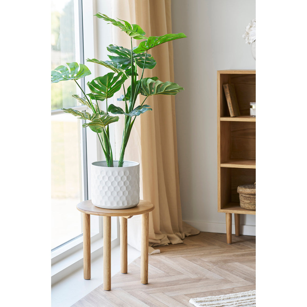 Cache-pot blanc nid d'abeille avec une grande plante verte posé sur une sellette en bois devant une fenêtre