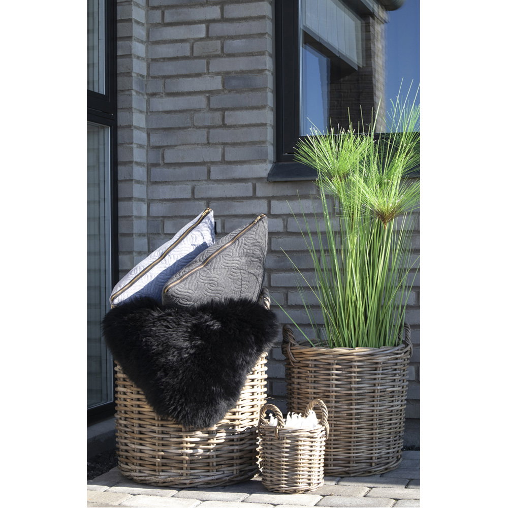 Trois cache-pots Caor en rotin kubu garnis de coussins d'une peau lainée noire et d'herbes hautes devant une façade grise