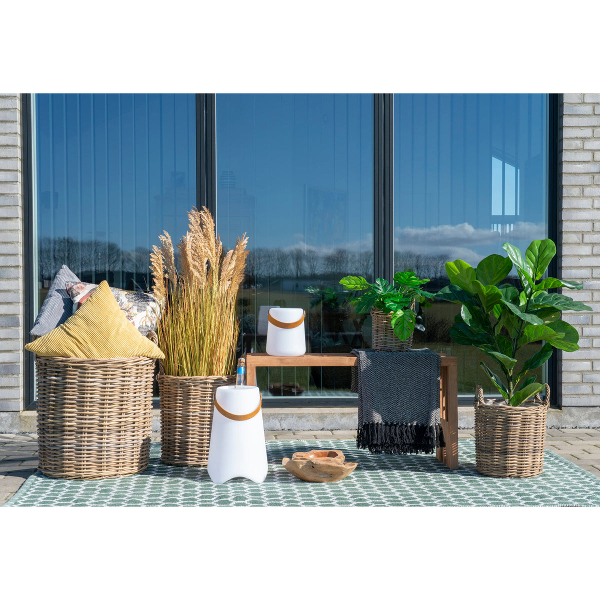 Quatre cache-pots Caor en rotin kubu garnis de coussins de graminées et de plantes vertes sur une terrasse devant une baie vitrée