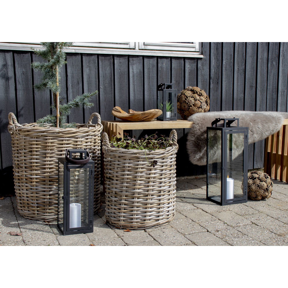 Les deux cache-pots Indo en rotin kubu sur une terrasse pavée entre des lanternes noires et un banc en bois
