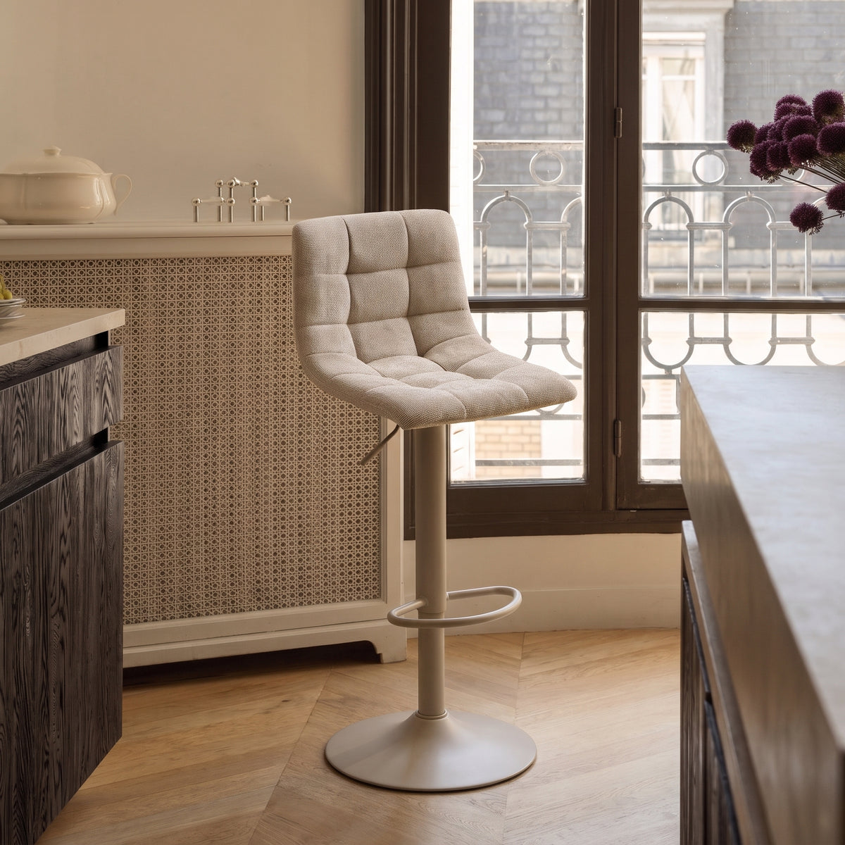Chaise de bar Middelfart en tissu gris devant un plan de travail de cuisine et une fenêtre, sur un parquet en chevrons