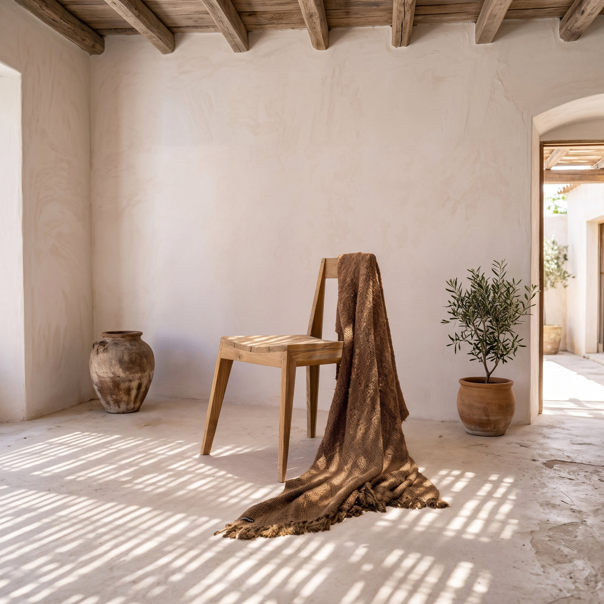 Chaise Paxi en teck naturel drapée d'un plaid brun dans une pièce aux murs chaulés