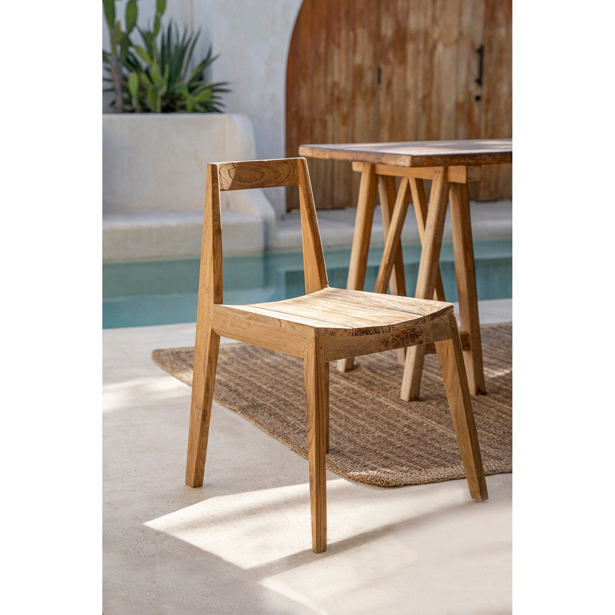 Chaise Paxi en teck naturel de trois quarts devant une table en bois à tréteaux au bord d'une piscine