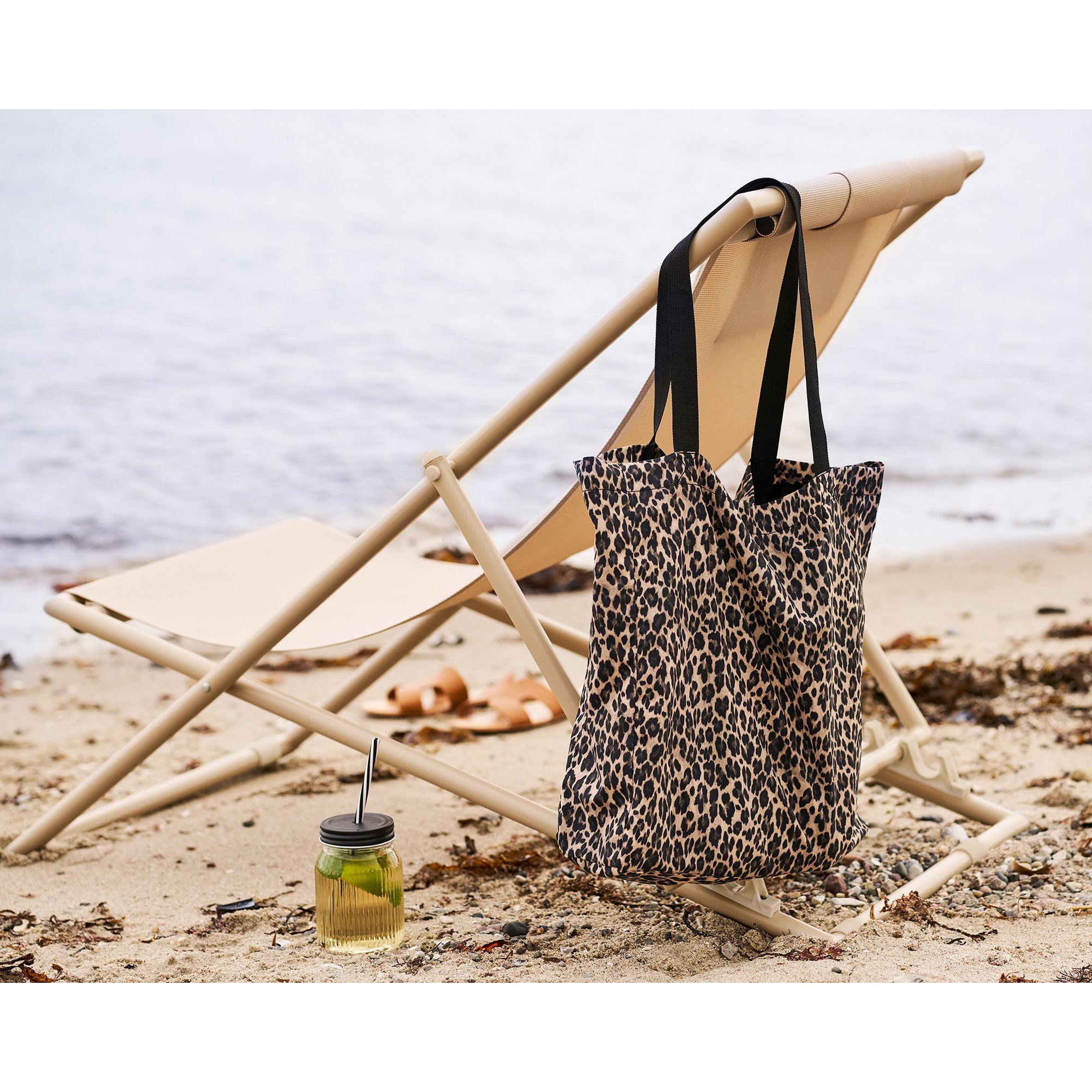 Chaise Curtis taupe repliée sur plage avec sac léopard accroché et boisson fraîche