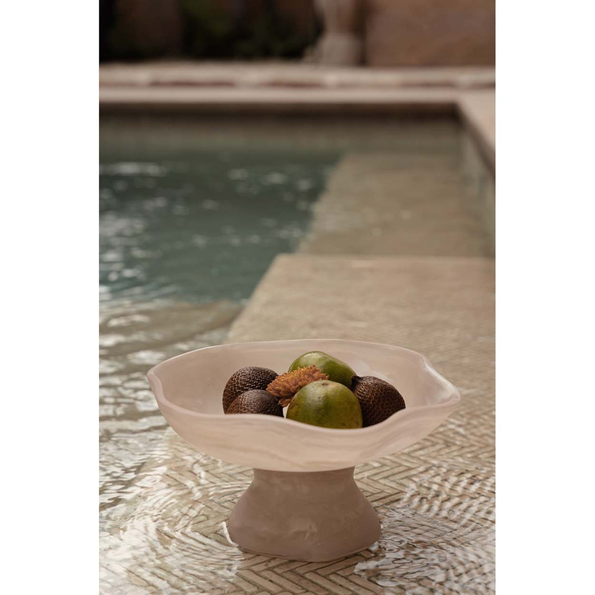 Coupe sur pied en résine rose marbrée garnie de fruits au bord d'une piscine sur un sol à chevrons