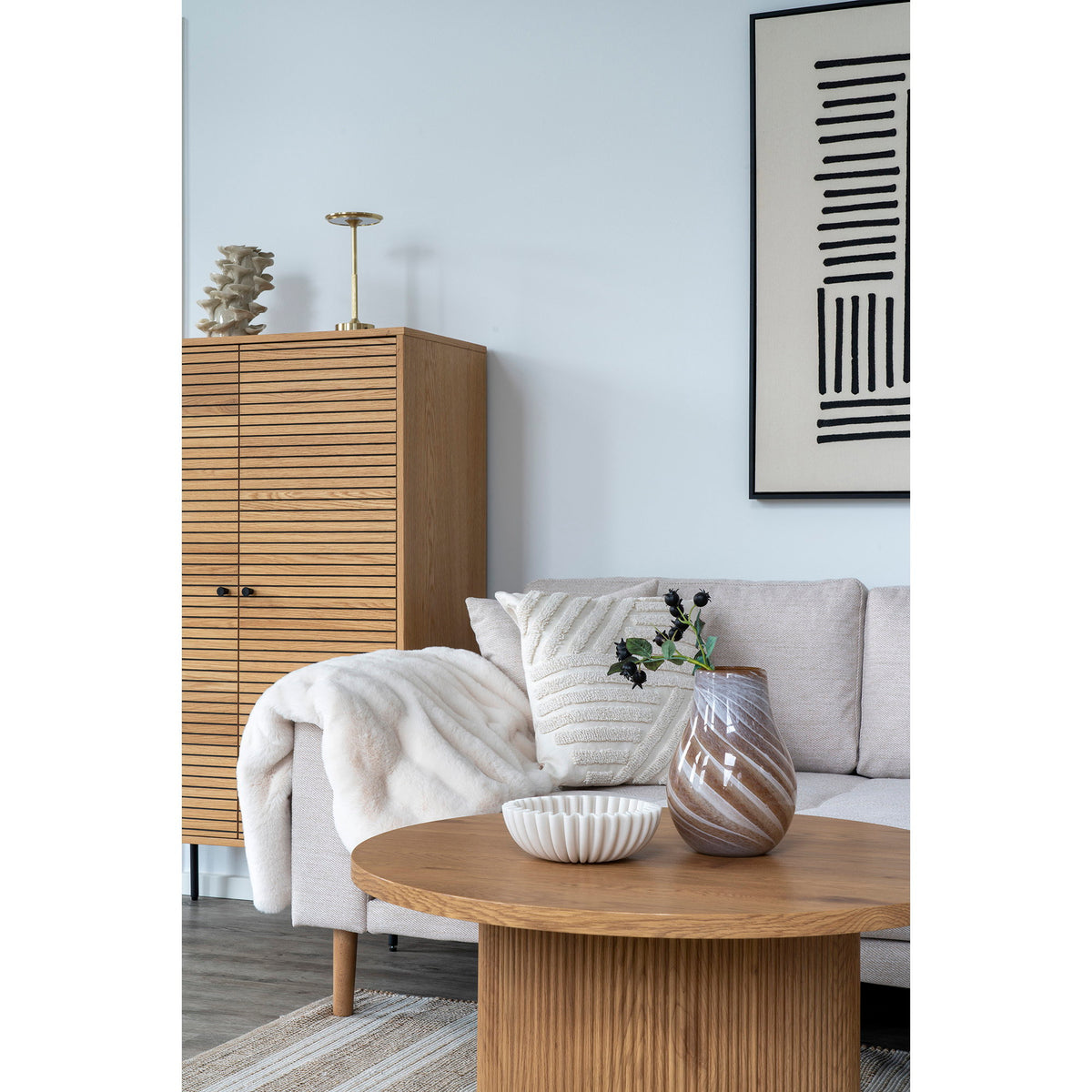 Coupe plissée beige sur une table basse ronde en bois à pied cannelé près d'un vase en verre tourbillonné devant un canapé clair