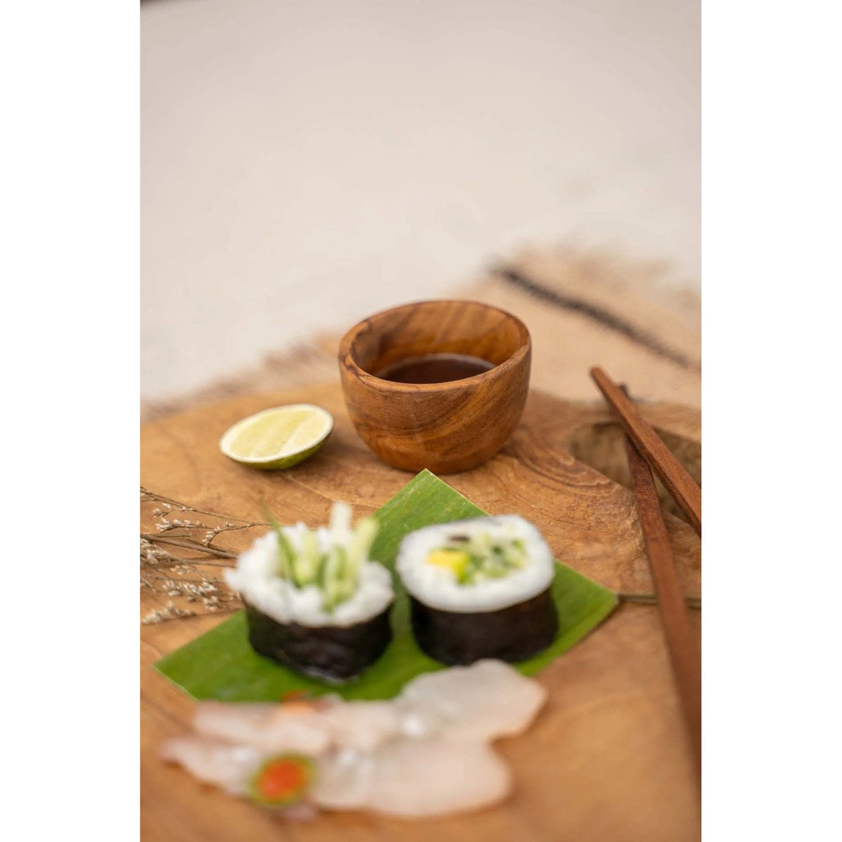 Coupelle en teck remplie de sauce soja sur une planche en bois près d'un demi-citron vert et de deux makis sur une feuille de bananier
