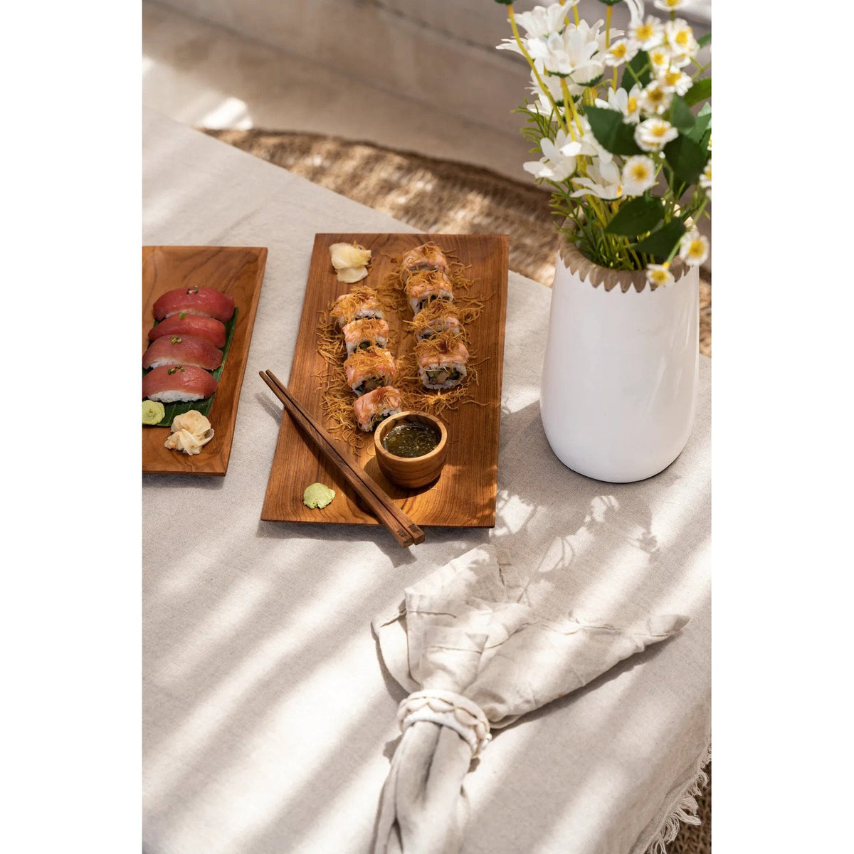 Table dressée pour des sushis avec une coupelle de sauce en teck sur un plateau en bois près d'un vase blanc garni de fleurs