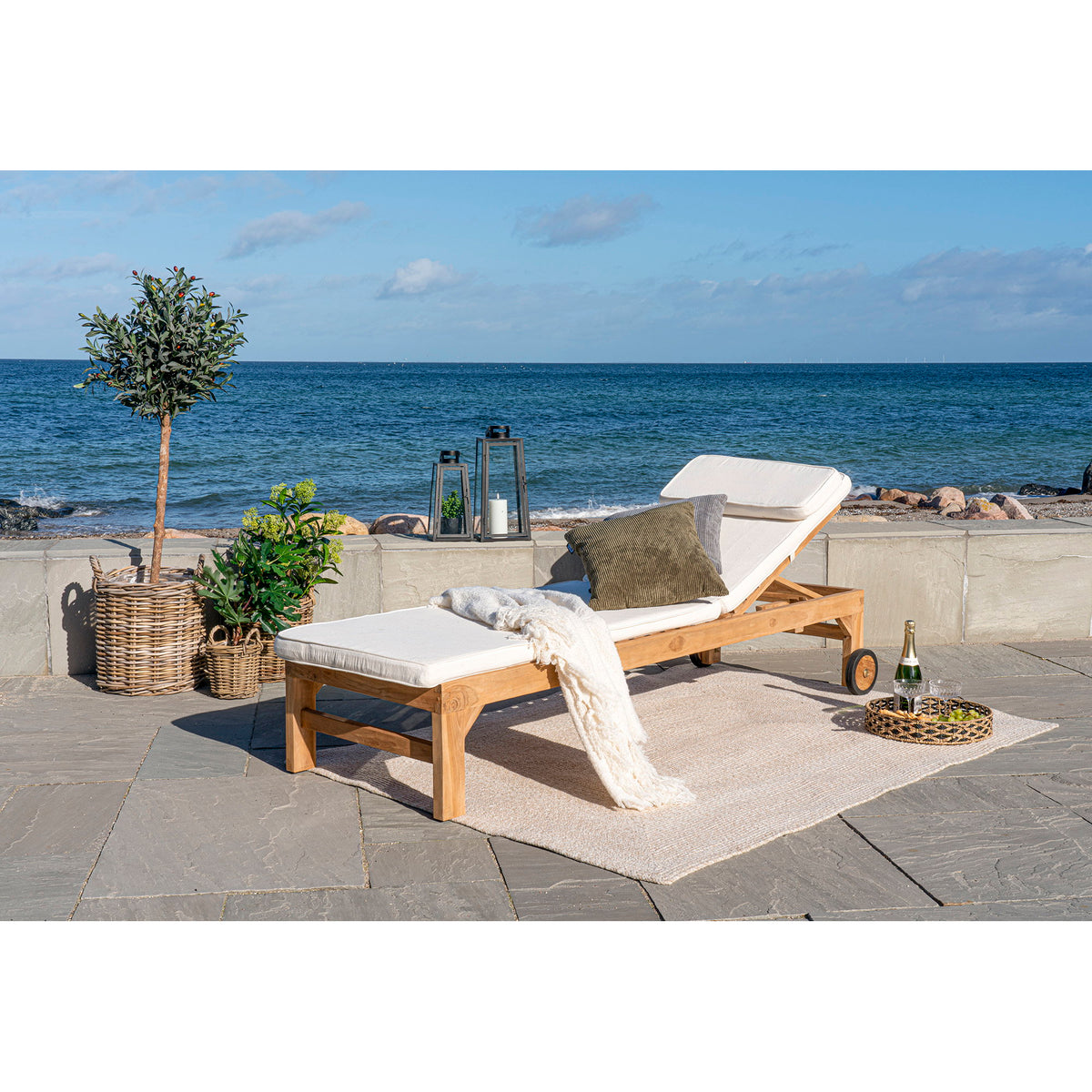 Coussin Andorra blanc sur un bain de soleil en teck sur une terrasse face à la mer avec un plaid et deux lanternes
