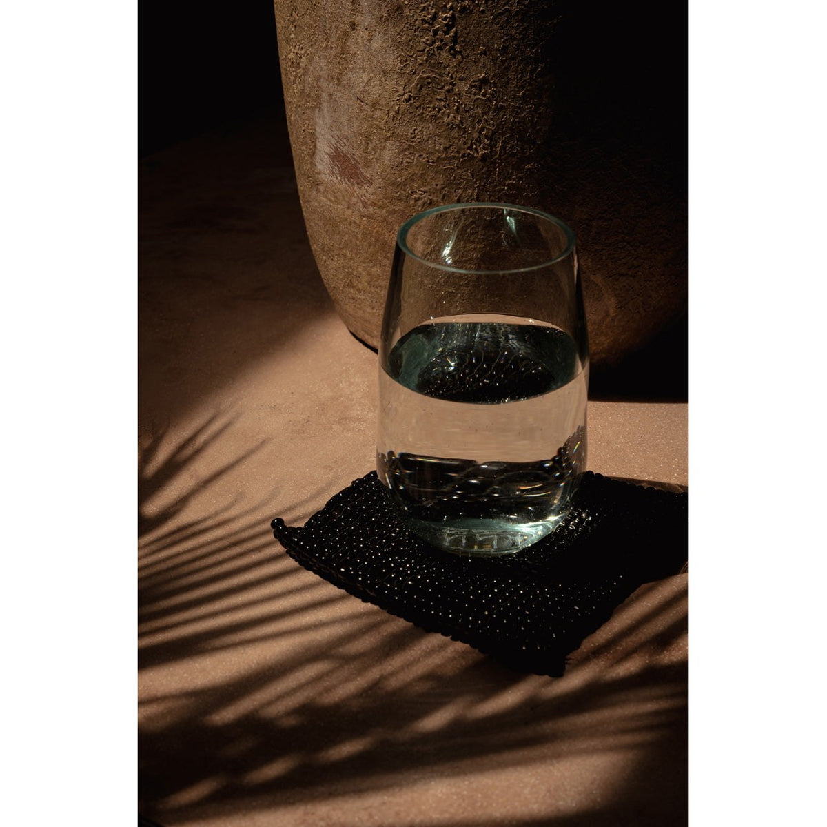 Dessous-de-verre carré en perles noires sous un gobelet d'eau à côté d'un grand pot en terre dans l'ombre d'une palme