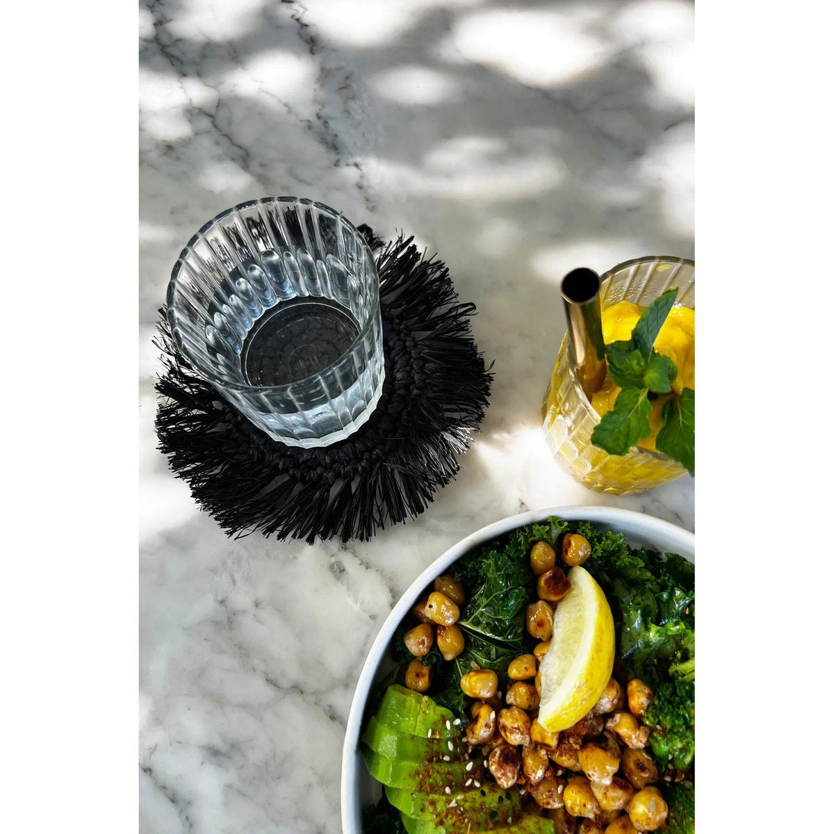 Verre d'eau strié posé sur le dessous-de-verre noir à frange sur une table en marbre blanc près d'une salade