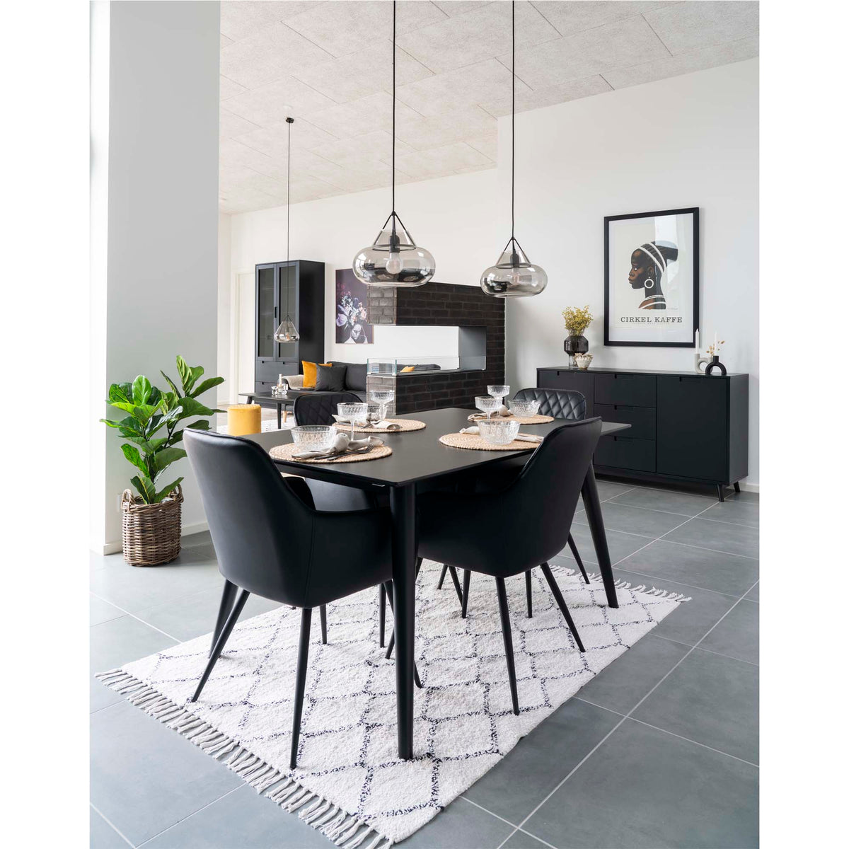 Quatre fauteuils Harbo simili cuir noir mat autour d'une table noire dressée sur un tapis blanc sous deux suspensions en verre fumé