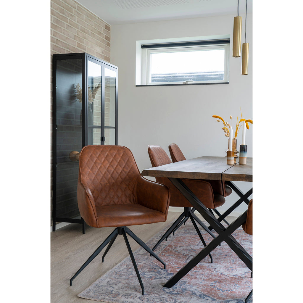 Gros plan fauteuil Harbo pivotant cuir vintage brun matelassé losanges autour table bois avec vitrine et mur briques