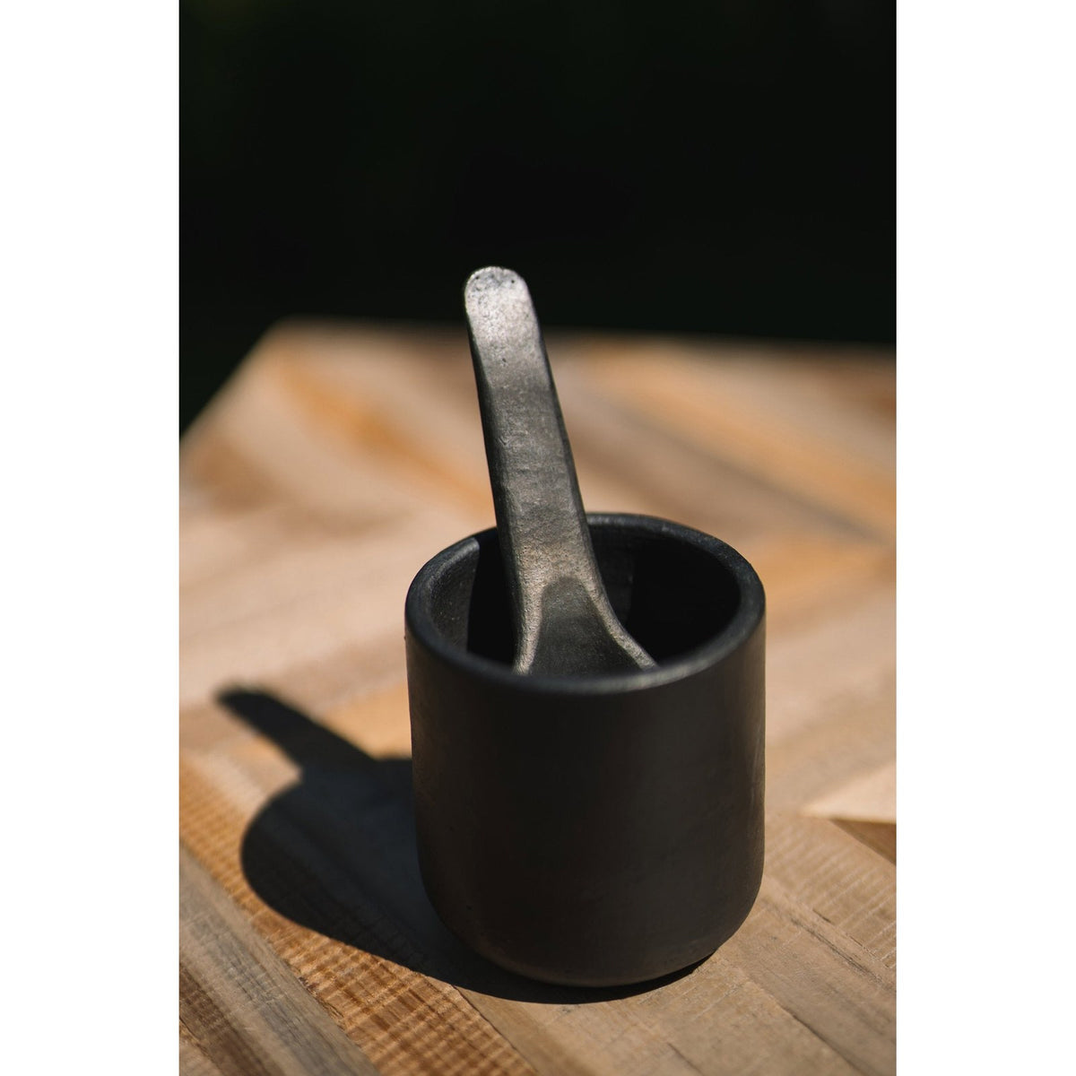 Gobelet en terre cuite noire avec une cuillère en métal sombre sur une planche en bois au soleil