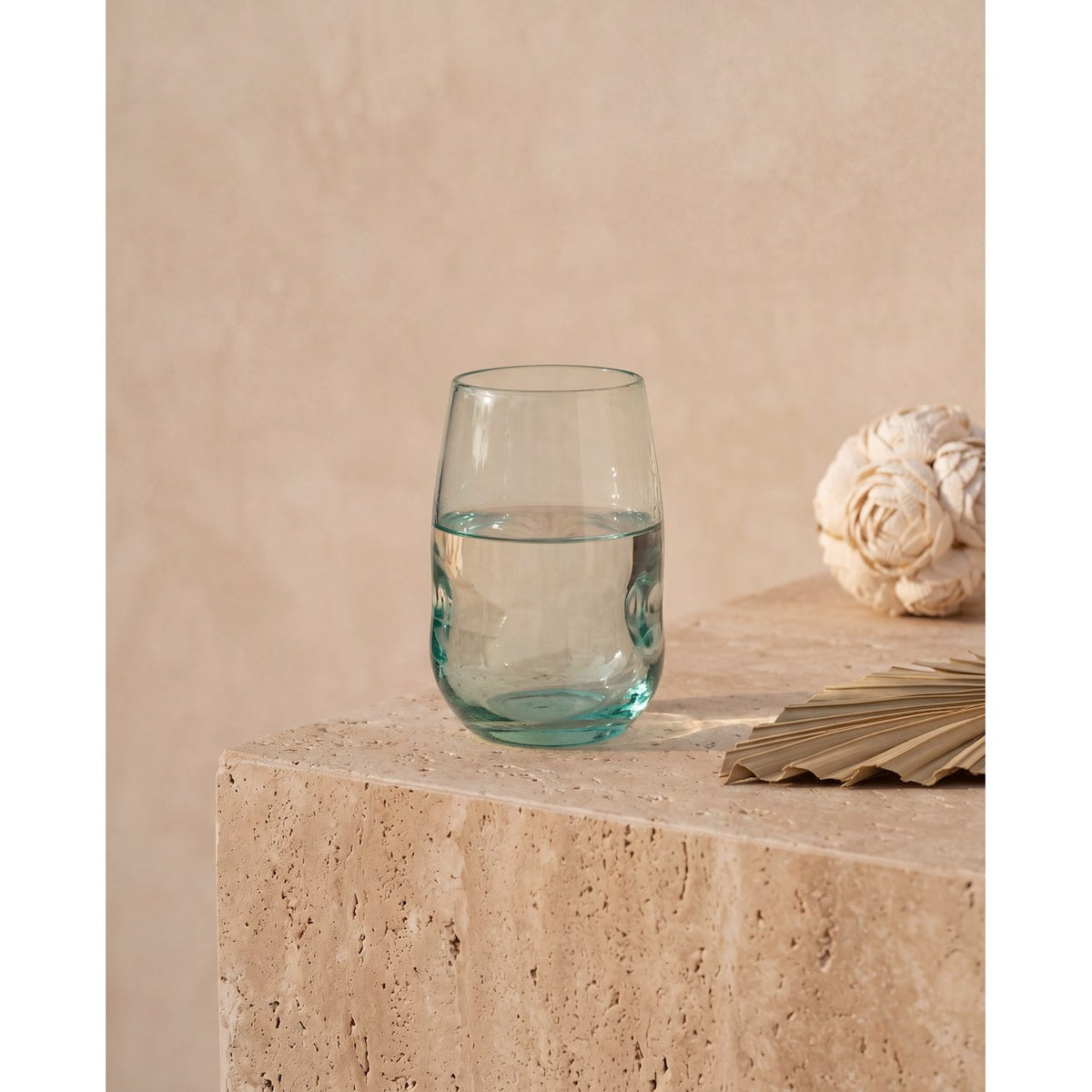 Gobelet galbé en verre recyclé bleuté rempli d'eau posé sur un bloc de pierre claire près d'une fleur séchée