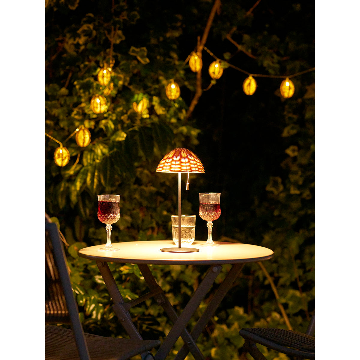 Guirlande Carlisle allumée dans les arbres en fond derrière une lampe de table et trois verres