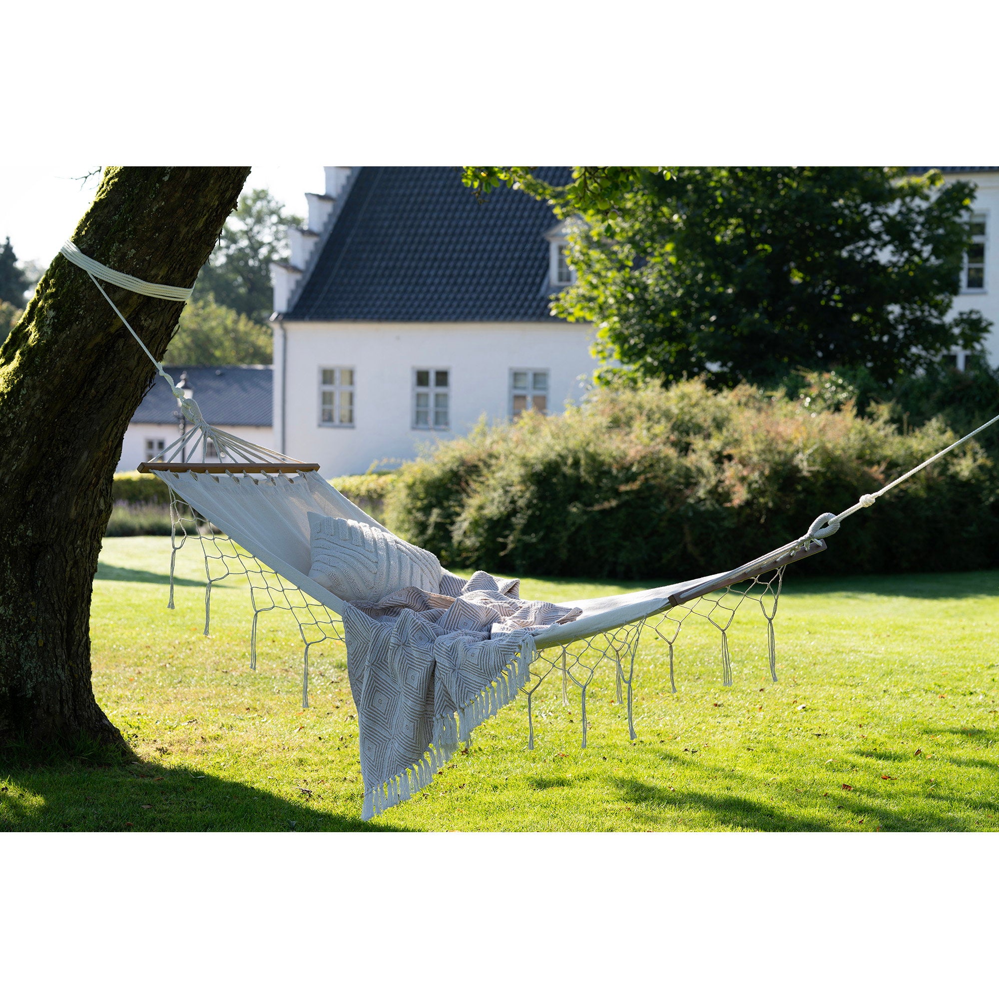 Hamac Edessa blanc cassé suspendu entre deux arbres avec coussins et plaid