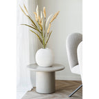 Herbe décorative artificielle beige sable dans un vase boule blanc sur une table d'appoint ronde près d'un fauteuil bouclé