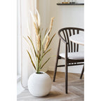 Herbe décorative artificielle beige sable dans un vase boule blanc strié posé au sol près d'une chaise en bois