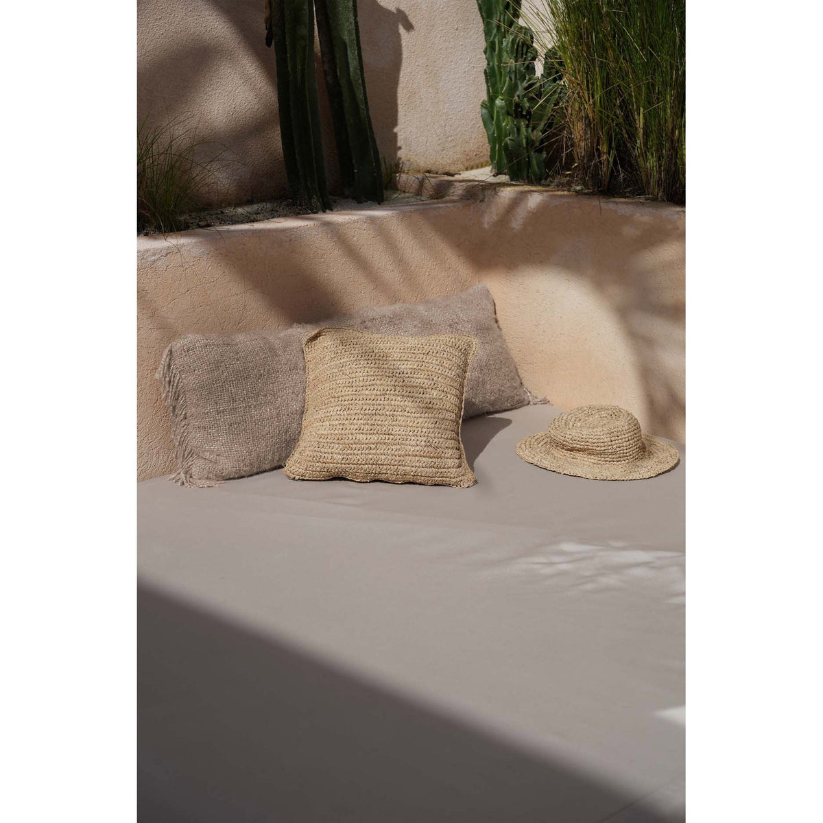 Coussin Flores en raphia sur une banquette de patio devant des cactus avec un chapeau de paille