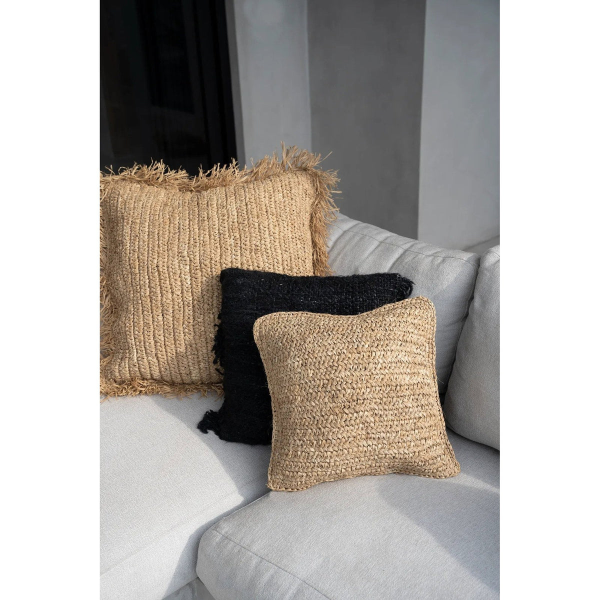 Coussin Flores en raphia au premier plan d'un coussin en raphia à franges et d'un coussin noir sur un canapé gris clair