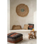 Coussin Flores en raphia entre un coussin à franges et un coussin tissé noir et orange sous un miroir en raphia