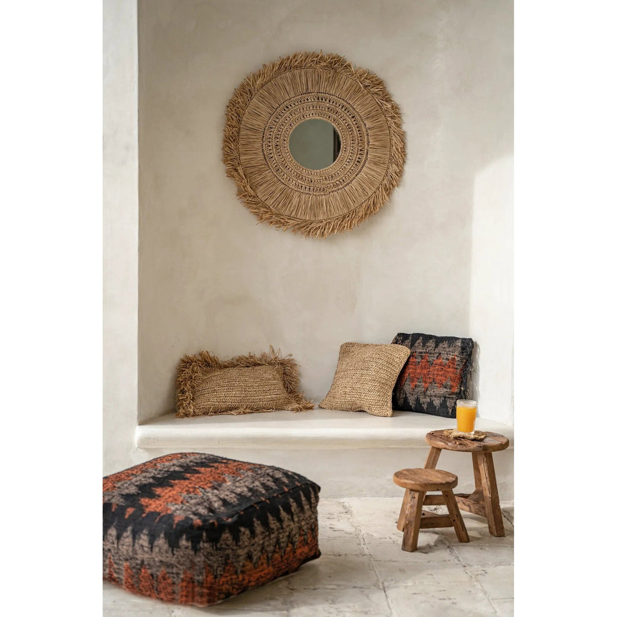 Coussin Flores en raphia entre un coussin à franges et un coussin tissé noir et orange sous un miroir en raphia