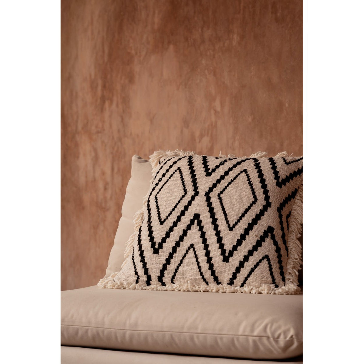 Housse de coussin Sahara Diamond de trois quarts sur une assise écrue devant un mur terracotta