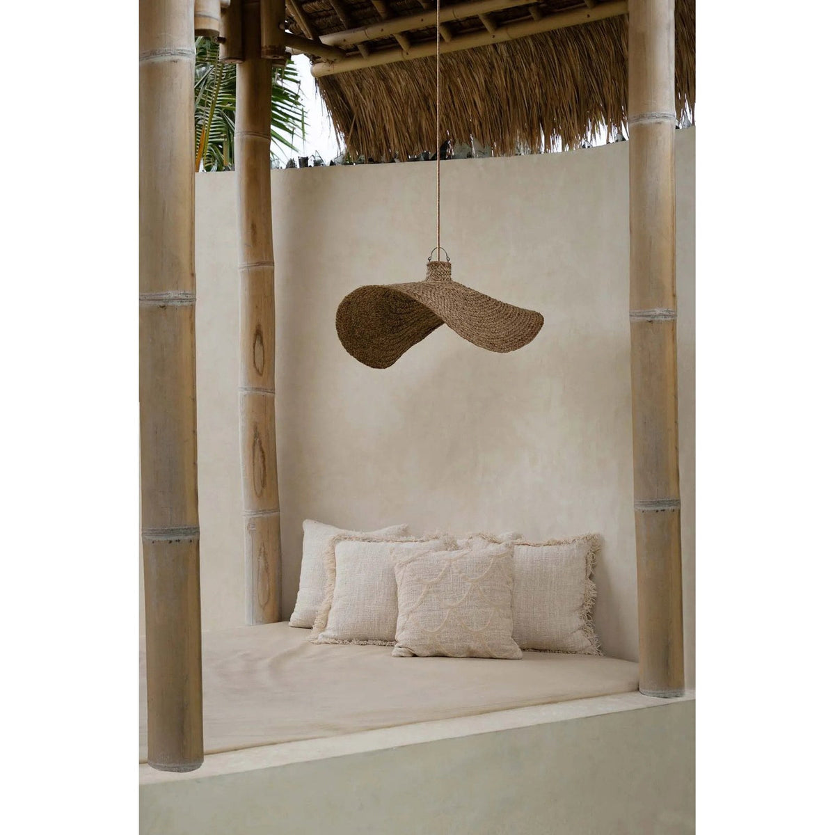 Coussin Sahara au premier plan d'une banquette sous une pergola en bambou avec des coussins frangés et une suspension en fibre