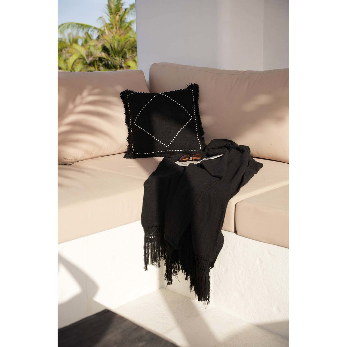 Jeté de lit Gypsy en coton noir posé sur une banquette beige à côté d'un coussin noir à surpiqûres blanches