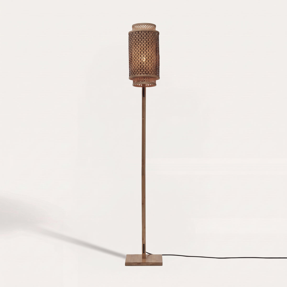 Lampadaire Bhutan allumé sur fond clair, abat-jour cylindrique en bambou tressé naturel et noir sur un pied en bambou