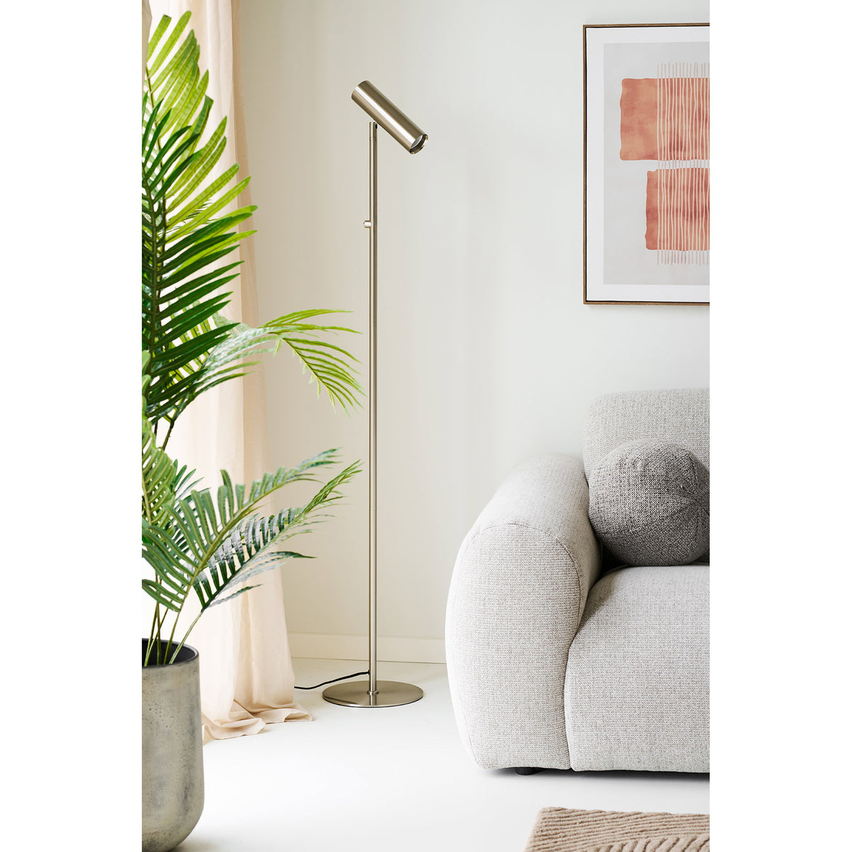 Lampadaire Paris finition acier brossé vu en entier entre un palmier en pot et un canapé gris clair sous un tableau terracotta