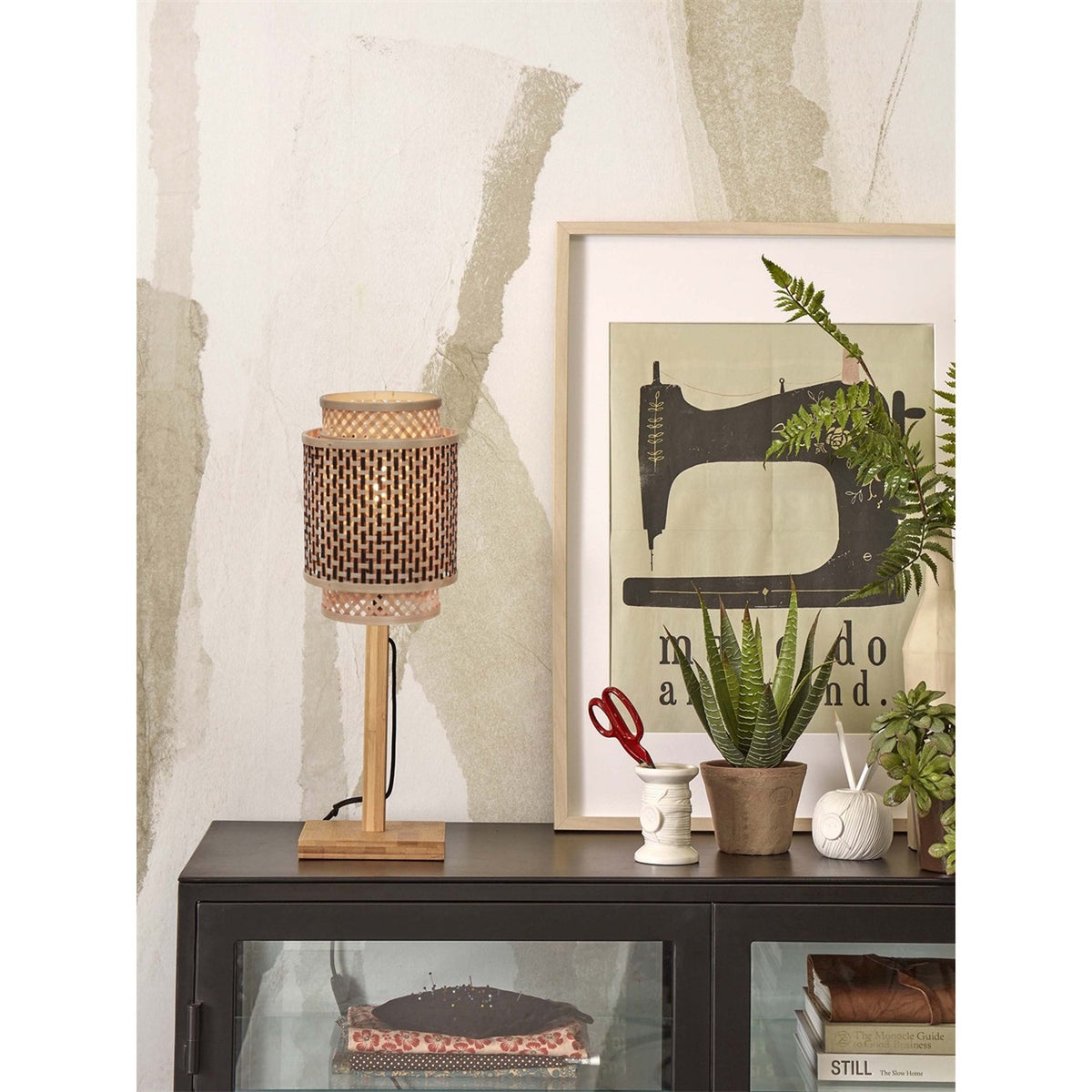 Lampe à poser Bhutan allumée sur un buffet vitré noir à côté d'une affiche encadrée et de plantes