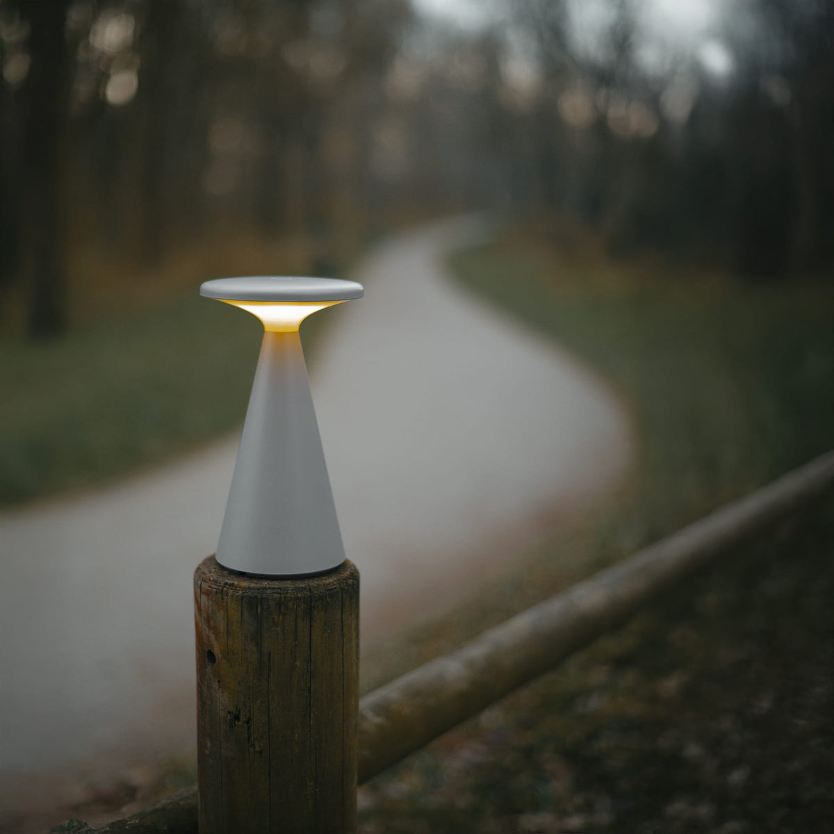 Lampe à poser Funghi grise allumée posée sur un poteau en bois au bord d'un chemin