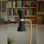 Abat-jour diabolo noir mat de la lampe Cornet avec sa bague et son bras dorés devant une bibliothèque